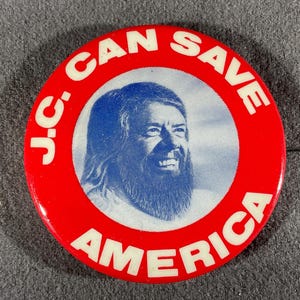 May include: A round, red button with white text that reads "J.C. CAN SAVE AMERICA." The center features a blue and white portrait of a man with long hair and a beard.