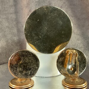 May include: Three gold-toned round earrings with a textured surface. The earrings are on a white surface. The back of one earring has the word "Vendome" printed on it.