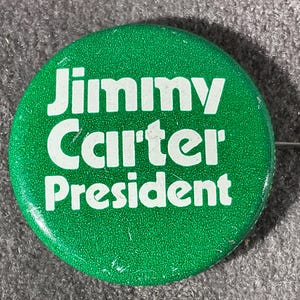 May include: A round, green button with the text "Jimmy Carter President" in white. The button has a pin on the back and is set against a gray, textured background. The button is a political campaign item.