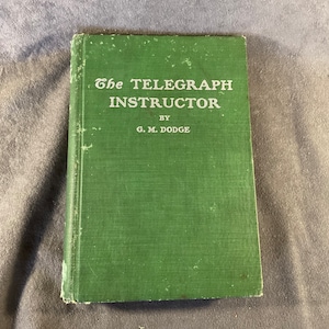 May include: A green book titled "The Telegraph Instructor" by G. M. Dodge.
