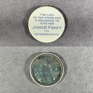 May include: A vintage campaign button with the text "THE LADY ON THE OTHER SIDE IS REQUESTED TO VOTE FOR JOHN D. FAHEY FOR REPRESENTATIVE" in blue on a white background. The button's reverse side is visible, showing a blue and gray design.