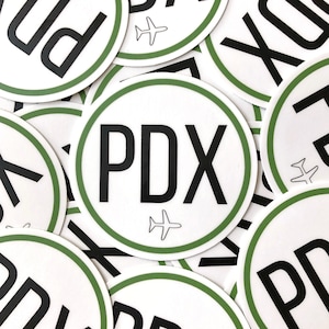 Op de afbeelding: Een close-up van verschillende ronde stickers met een groene rand en een witte achtergrond. De stickers tonen de tekst "PDX" in zwarte letters met een klein vliegtuigpictogram eronder.