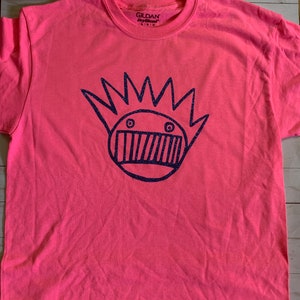 May include: A pink t-shirt with a blue glitter design of a cartoon face with a crown of spikes. The shirt has a Gildan DryBlend tag on the front.