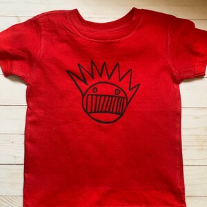 May include: Red short-sleeved t-shirt with a black graphic of a cartoon face with a spiky hairdo.