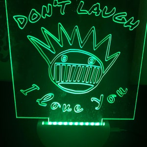 May include: Green LED light with a white base. The light features a clear acrylic panel with a black and white graphic of a cartoon character with the text "Don't Laugh I Love You".
