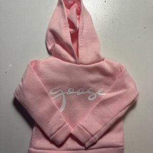 May include: A light pink hooded sweatshirt with the word "Goose" in white script across the chest. The sweatshirt has long sleeves and a ribbed hem and cuffs. The hood is lined and has a drawstring.