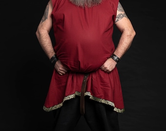May include: A man wearing a red tunic with a gold trim, black pants, and a green hat with red feathers. He has a long gray beard and is standing with his hands on his hips.