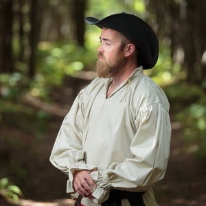 May include: A man wearing a white shirt with a black hat and red trousers stands in a forest. He is wearing a brown leather belt with a silver buckle and a sword.