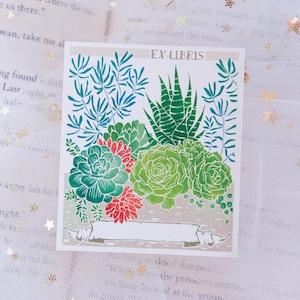 May include: A bookplate with the text "EX-LIBRIS" printed in black lettering. The bookplate features a colorful illustration of a variety of succulents in shades of green, red, and brown.