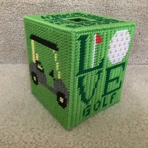 May include: Green plastic canvas tissue box cover with a golf theme. The box features a golf cart, a golf ball, and the words "LOVE GOLF" in green and white. The top has a square opening for tissue dispensing.