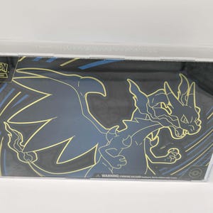 May include: A sealed Pokémon Trading Card Game box featuring a black and gold design with a stylized Charizard illustration. The box is encased in a clear plastic protector. The Pokémon logo is visible in the upper left corner.