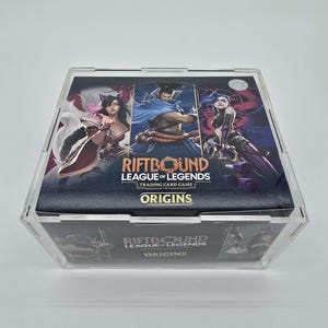 May include: A clear, acrylic display case holding a box of "RIFTBOUND LEAGUE OF LEGENDS TRADING CARD GAME ORIGINS" cards. The card box features three illustrated characters and the game's title. The case is on a white surface.