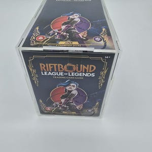 May include: A clear acrylic display case containing a "Riftbound League of Legends" trading card game box. The box features artwork of a character named Jinx and the text "4 Champion Decks". The box has a dark blue and gold colour scheme.