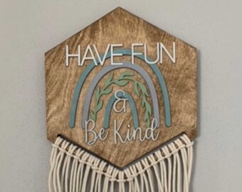 Puede incluir: Una pared hexagonal de madera con un diseño de arcoíris y el texto "Have Fun & Be Kind". La pared tiene una franja de macramé blanco.