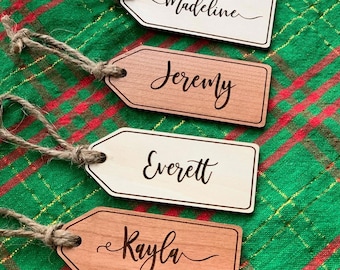 Puede incluir: Cuatro etiquetas de regalo de madera con un cordel atado. Las etiquetas tienen forma de rectángulo con esquinas redondeadas. Los nombres "Madeline", "Jeremy", "Everett" y "Kayla" están grabados en las etiquetas.