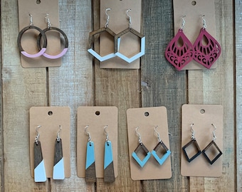 May include: Assortment of eight pairs of wooden earrings on display cards. The earrings are in various shapes and colors, including geometric shapes, circles, and floral designs. The earrings are painted in shades of pink, blue, black, and white.