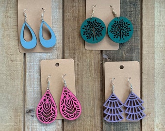 May include: Four pairs of wooden earrings on brown card stock. The earrings are in the shapes of teardrops, trees, and geometric designs. The earrings are painted in shades of blue, green, pink, and purple.