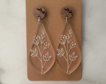 May include: A pair of clear acrylic dangle earrings with a white floral design. The earrings have wooden tops with a small floral design.