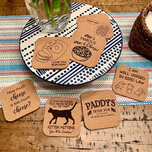 May include: Six cork coasters with different designs and sayings. The coasters are on a wooden table with a pink flower arrangement in a glass vase.
