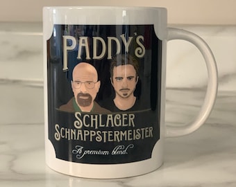 Puede incluir: Taza de cerámica blanca con un diseño negro y dorado. El diseño presenta dos retratos de dibujos animados de hombres con el texto "Paddy's Schlager Schnappsmeister A premium blend."