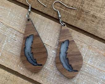 Puede incluir: Un par de pendientes de madera con forma de lágrima. Cada pendiente presenta un diseño de pluma plateada con un acabado brillante.