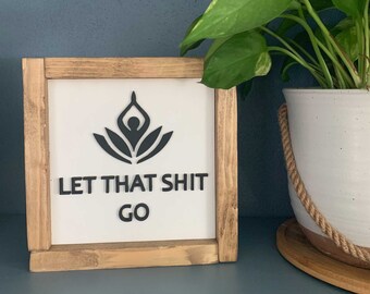 Puede incluir: Un letrero con marco de madera y fondo blanco. El letrero presenta una silueta negra de una persona en una postura de yoga con un diseño floral. El texto "LET THAT SHIT GO" está escrito en letras negras debajo del diseño.
