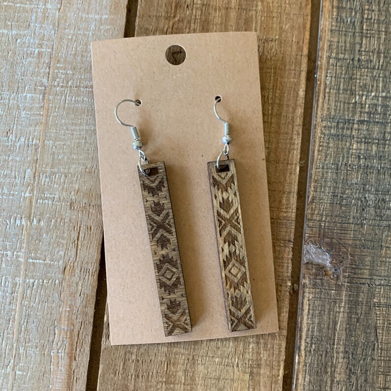 May include: A pair of wooden dangle earrings with a geometric design. The earrings are rectangular with a detailed pattern etched into the wood. They hang from silver-colored fish hook ear wires. The earrings are displayed on a brown card.