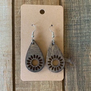 May include: A pair of wooden teardrop earrings with a sunflower design. The earrings are dark brown with silver-colored hooks and are displayed on a brown card.