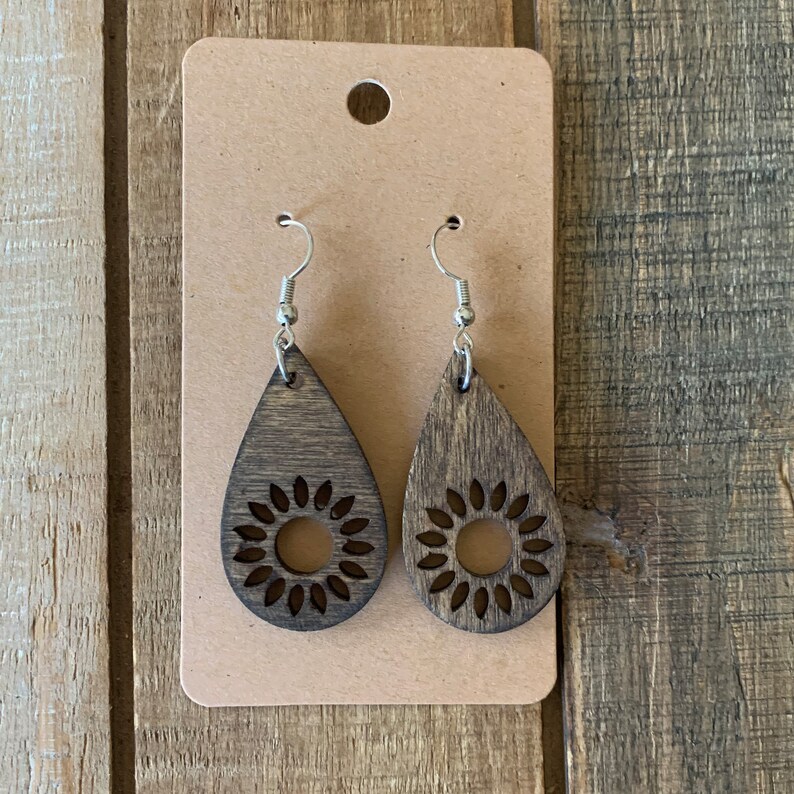May include: A pair of wooden teardrop earrings with a sunflower design. The earrings are dark brown with silver-colored hooks and are displayed on a brown card.