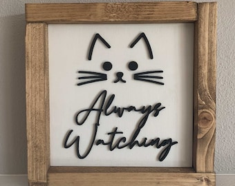 Puede incluir: Un letrero de madera con fondo blanco y letras negras. El letrero presenta una cara de gato estilizada con el texto "Always Watching".