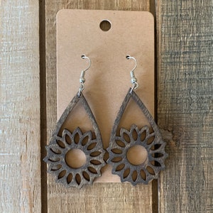 May include: Wooden teardrop earrings with a sunflower design. The earrings are made of dark brown wood and feature a cut-out sunflower in the center. The earrings hang from silver-colored fish hook ear wires. The earrings are displayed on a brown card.