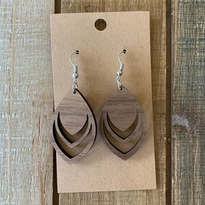 May include: A pair of wooden teardrop earrings with a layered chevron design. The earrings are a medium brown color and hang from silver-colored fish hook ear wires. The earrings are displayed on a brown card with a hole at the top.
