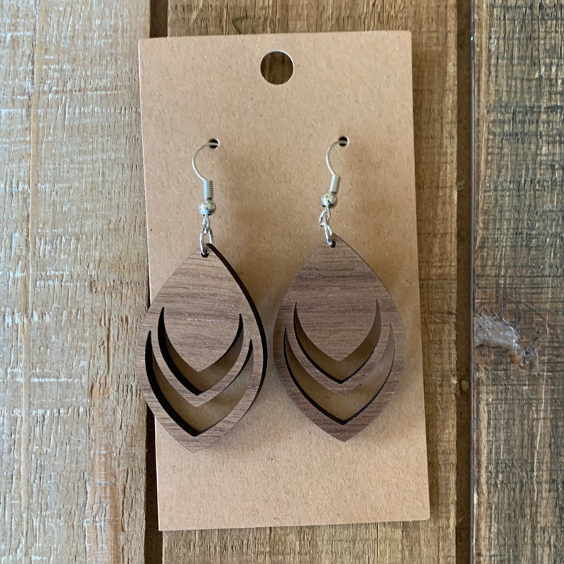 May include: A pair of wooden teardrop earrings with a layered chevron design. The earrings are a medium brown color and hang from silver-colored fish hook ear wires. The earrings are displayed on a brown card with a hole at the top.
