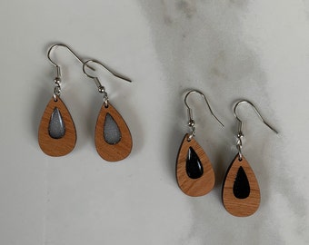 Puede incluir: Un par de pendientes de madera en forma de lágrima con ganchos plateados. Un par tiene una inserción de resina transparente con purpurina plateada, y el otro par tiene una inserción de resina negra.