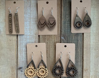 Puede incluir: Una colección de pendientes de madera exhibidos en tarjetas marrones. Los pendientes presentan varios diseños, incluyendo patrones geométricos, gotas superpuestas y recortes florales. Los tonos de madera varían de marrón claro a oscuro, con ganchos plateados.