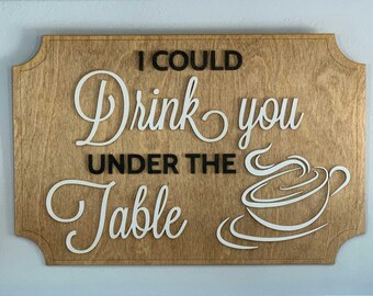Puede incluir: Un letrero de madera con el texto "I could drink you under the table" en letras negras y una ilustración de una taza de café blanca.