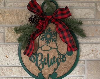 Puede incluir: Un colgador de puerta de madera con forma de gnomo con un gnomo pintado de verde, un lazo de cuadros rojo y negro y la palabra "Believe" escrita en verde.