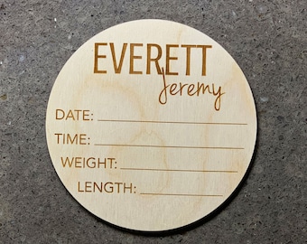 Puede incluir: Un disco redondo de madera clara con los nombres "EVERETT" y "jeremy" grabados en una fuente elegante. Debajo, se proporcionan líneas para registrar la fecha, la hora, el peso y la longitud. Un recuerdo para un recién nacido.