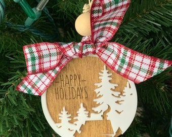 Puede incluir: Un adorno navideño de madera con una silueta blanca de pinos y el texto "Happy Holidays". El adorno está decorado con una cinta de cuadros rojos y verdes.