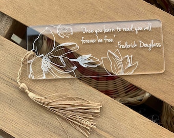 Puede incluir: Un marcapáginas de acrílico transparente con un diseño floral blanco y la cita "Una vez que aprendas a leer, serás libre para siempre. -Frederick Douglass". El marcapáginas tiene un borla.