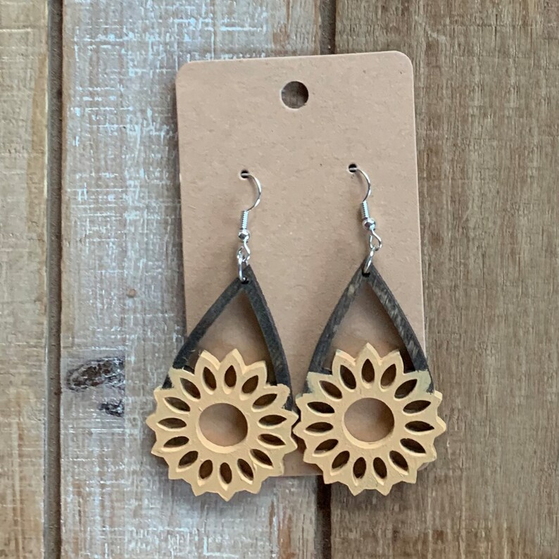 May include: Wooden sunflower earrings with silver hooks. The earrings feature a teardrop-shaped frame in dark brown, with a light brown sunflower design in the center. The earrings are displayed on a brown card.