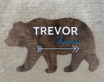 Puede incluir: Silueta de oso de madera con los nombres "Trevor Thomas" y una flecha gris con detalles azules.
