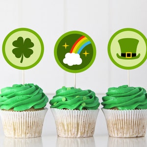 Op de afbeelding: Drie cupcakes met groene frosting en decoratieve toppers. De toppers tonen een klavertjevier, een regenboog met een wolk en een groene kabouterhoed. De cupcakes zitten in witte papieren vormpjes, perfect voor St. Patrick's Day.