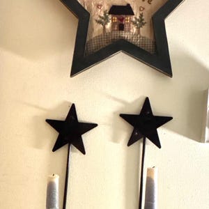 May include: A black star-shaped wall decor with the text "Home of the brave" and a framed picture of a house. Below are two black star-shaped candle holders with gray candles.