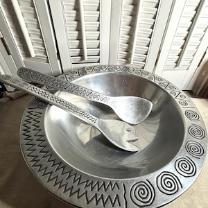 May include: A silver-colored salad bowl with matching serving utensils. The bowl and utensils have a decorative etched design around the rim and on the handles. The bowl is round and has a wide rim. The serving utensils include a spoon and a fork.