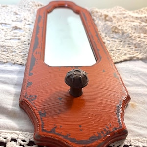 May include: A small, rectangular mirror with a distressed orange wooden frame and a decorative knob. The frame has gray accents, and the mirror is mounted on a decorative wooden backing. The overall design has a vintage, rustic aesthetic.