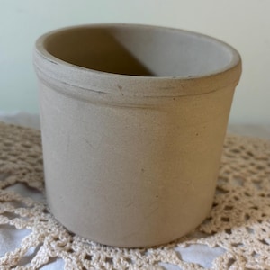 May include: A small, beige ceramic crock with a rounded bottom and a slightly flared rim. The crock is sitting on a white crocheted doily.