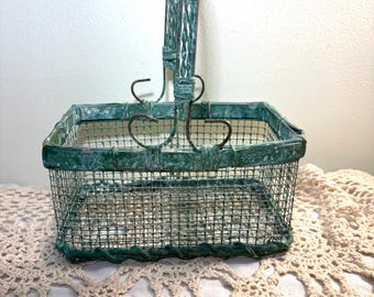 May include: A rectangular wire basket with a handle, painted in a distressed teal green. The basket has a square shape and is made of metal wire. The handle is arched and attached to the basket with decorative metal swirls.