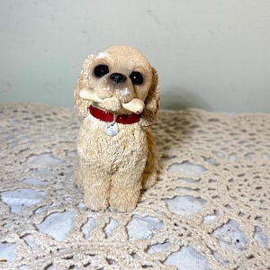 May include: A tan-colored dog figurine with black eyes and a red collar, holding a bone in its mouth. The dog has floppy ears and a small tag on its collar. The figurine is sitting on a beige, patterned surface.
