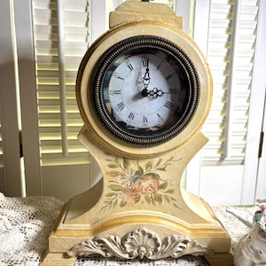 May include: A cream-colored mantel clock with a round face featuring black Roman numerals and ornate hands. The base and frame are decorated with painted floral motifs and a carved shell-like detail at the bottom, topped with a dark metal finial.